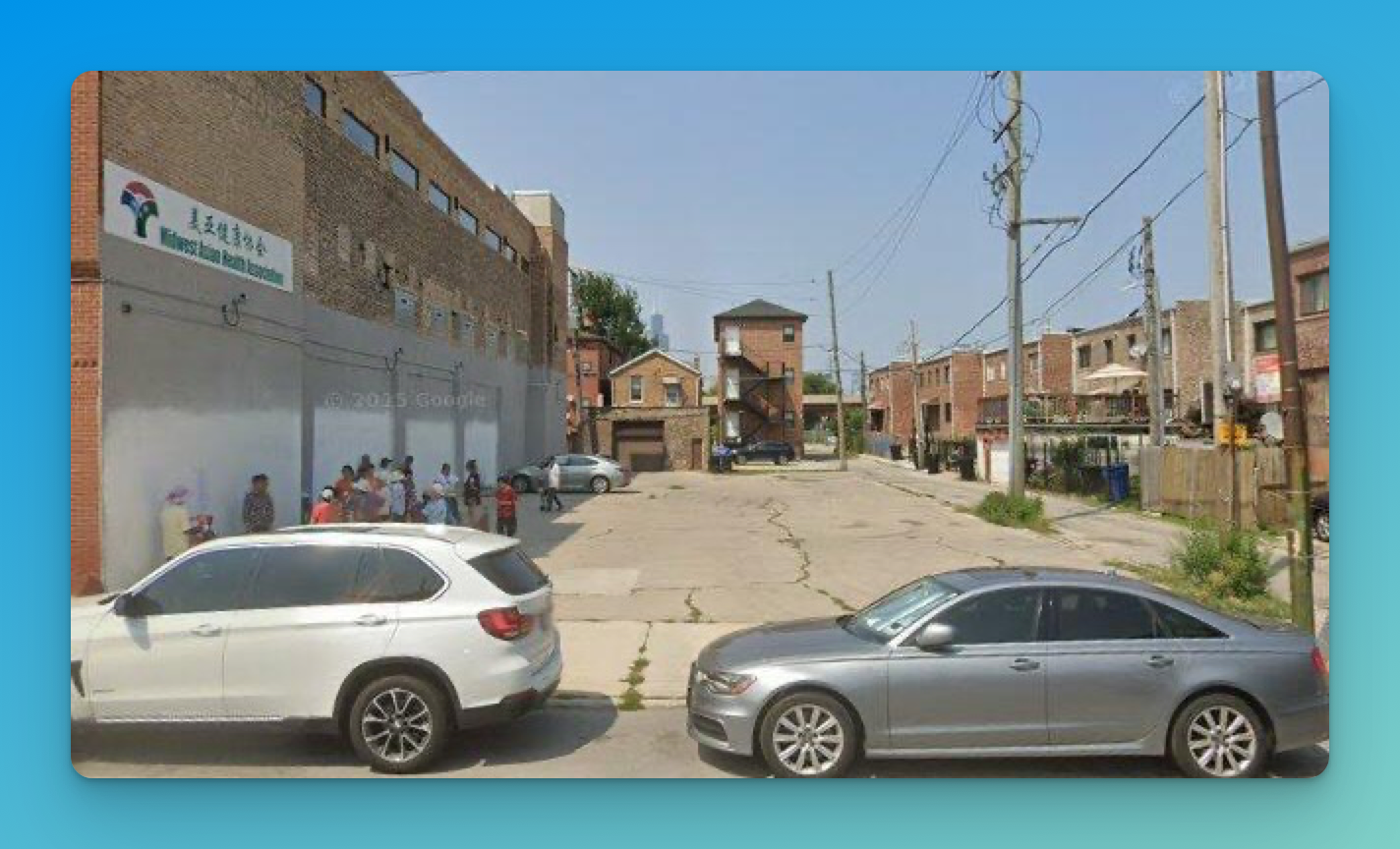 Google Street View of the now-vacant lot in Armour Square, just north of Chinatown, seen from 26th Street. The Midwest Asian Health Association building is on the left and cars are parked along the curb in front of the open parcel.